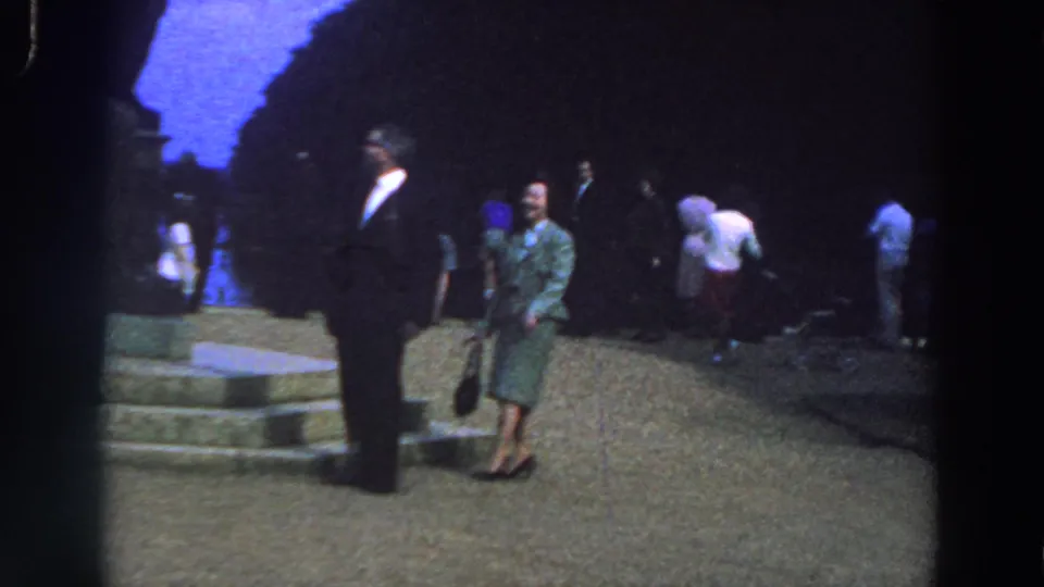 1961 Ireland home-movie frame with a woman in a green dress turning toward camera near a suited man, stone steps, and background visitors.