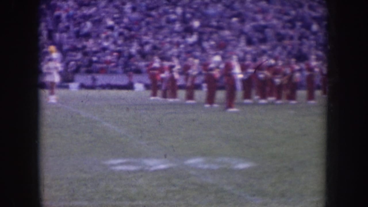 Nebraska 1957 home-movie frame showing a red-uniformed marching band on a football field with spectators filling the stands behind them.