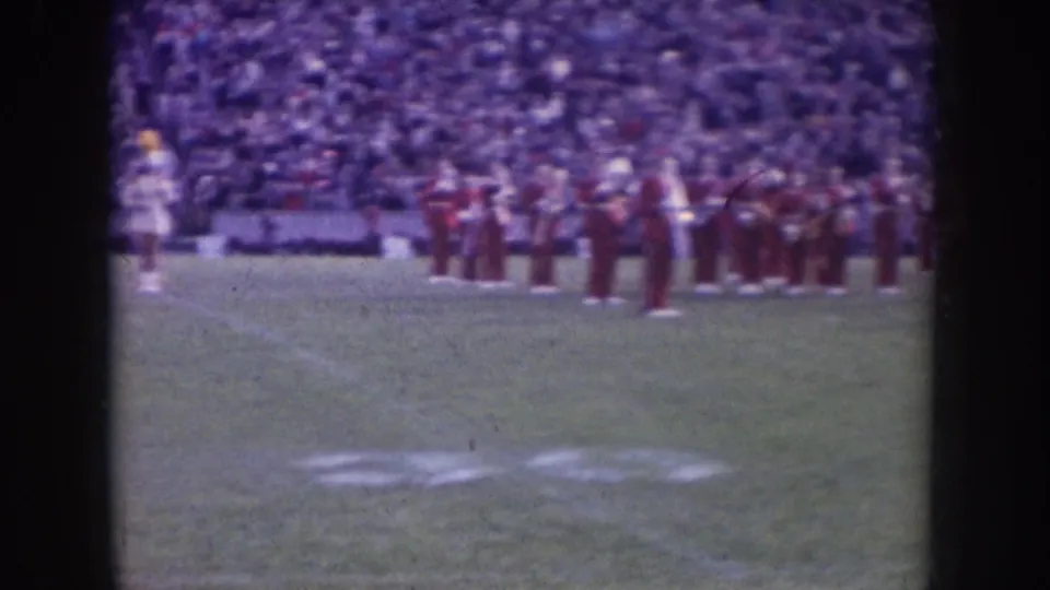 Nebraska 1957 home-movie frame showing a red-uniformed marching band on a football field with spectators filling the stands behind them.