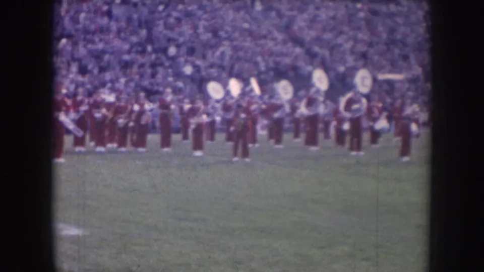 Nebraska 1957 frame with marching band members in red uniforms, including brass and percussion players, standing before packed stadium seating.