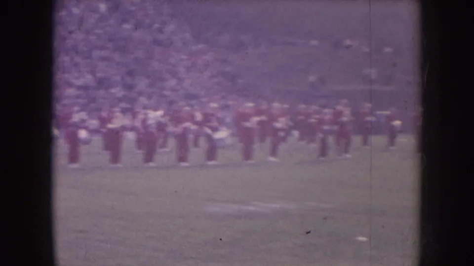 Nebraska 1957 home-movie frame with a hazy purple cast, showing a red-uniformed band lined across a football field.