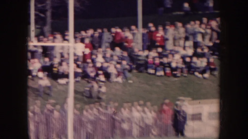 Nebraska 1957 frame showing goal posts, sideline spectators, crowded seating, and band members near a football field.