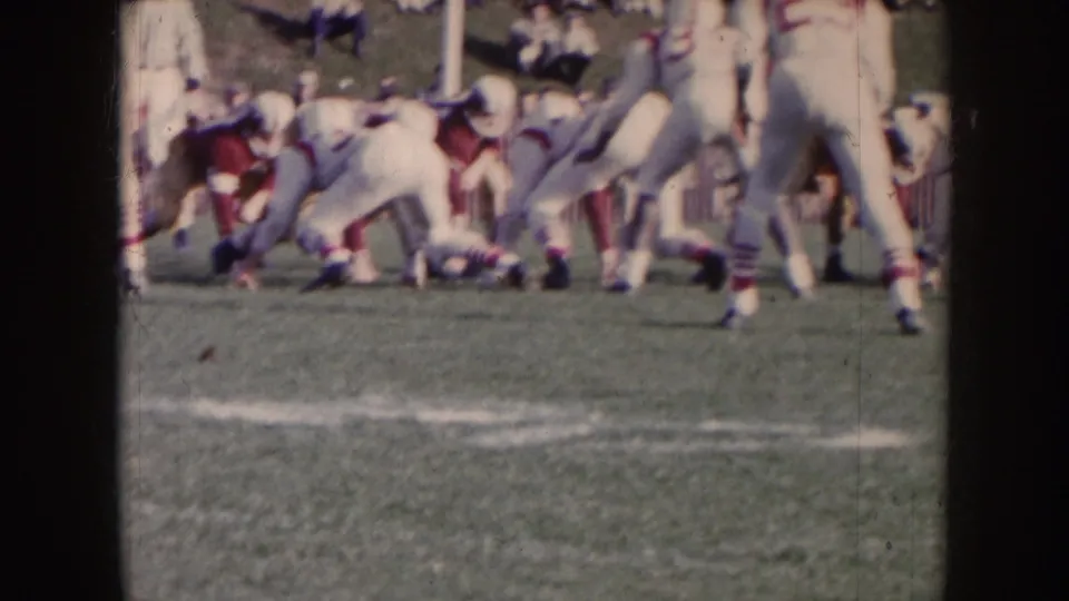 1957 Nebraska home-movie frame shows white-uniformed players and red-jerseyed opponents clustered at the line during football action.