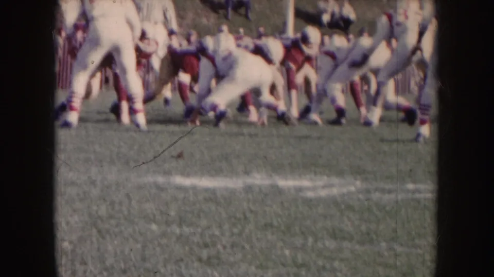 1957 Nebraska 8mm frame shows red and white football players in motion on a grass field, with soft focus and visible film wear.