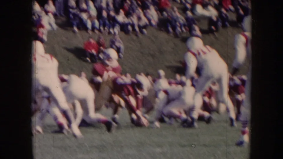 1957 Nebraska home-movie frame showing several football players collapsing into a tackle with spectators blurred in the background.