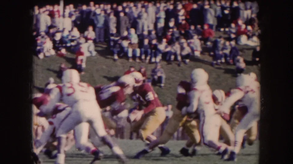 1957 Nebraska home-movie frame showing football action at the line with maroon and white players and a standing crowd behind them.