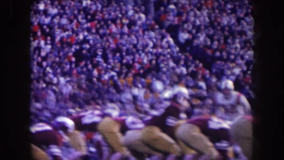 1957 Nebraska 8mm frame of helmeted football players at the line of scrimmage with spectators filling the hillside behind them.