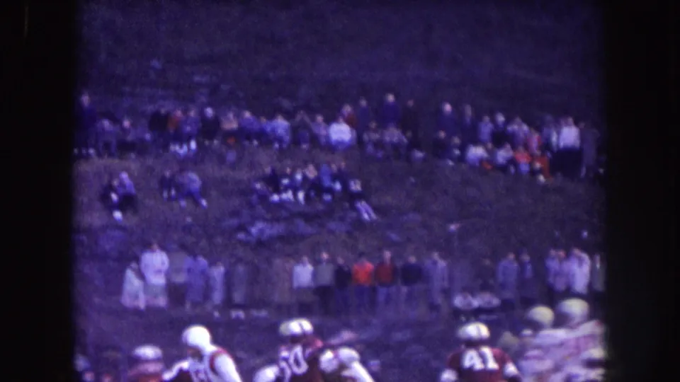 1957 Nebraska 8mm frame with football players at the lower edge and a long row of spectators across the grassy hillside.