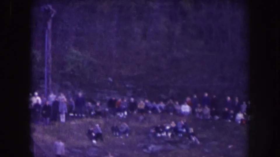Nebraska 1957 home-movie frame showing a distant row of spectators near a sports field, with heavy grain and soft focus.