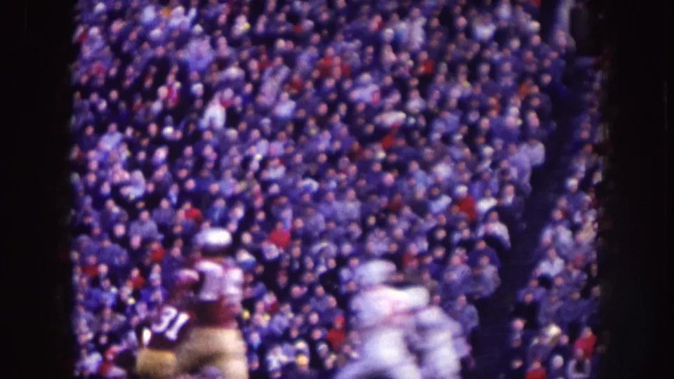 Nebraska 1957 home-movie frame showing helmeted field-sport players in front of packed stadium spectators.