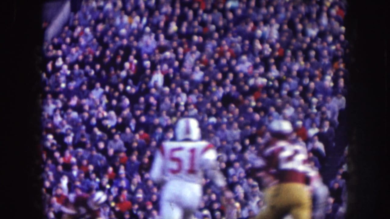 Football players run in front of a packed stadium crowd.