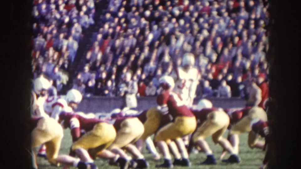 Players in white and maroon uniforms line up on a sunny football field, with crowded stands visible in the background.