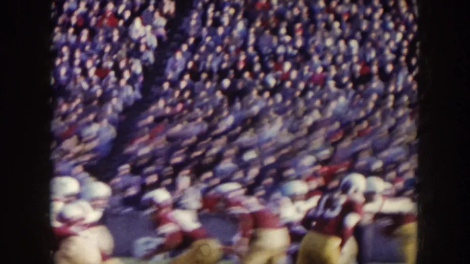 A fast-moving football play appears blurred in 8mm footage, with white and maroon uniforms and crowded stands behind the action.