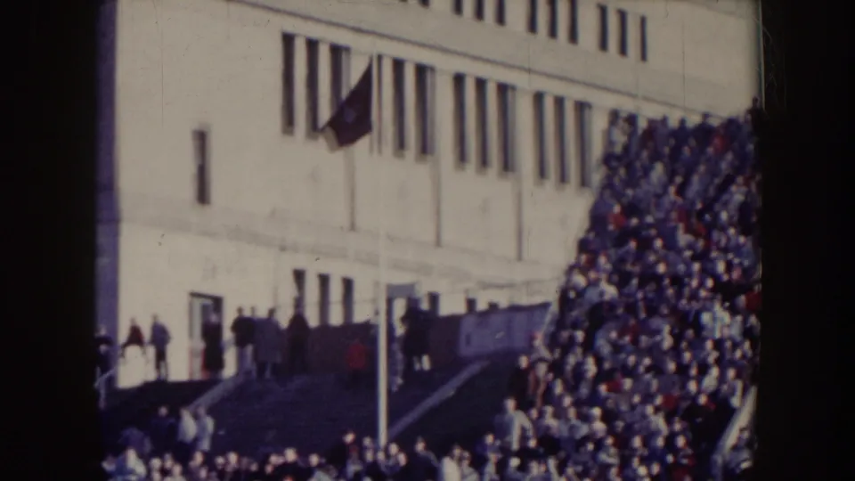 A stadium wall, dark flag on a pole, and packed Nebraska football stands appear in a handheld 1957 home-movie frame.