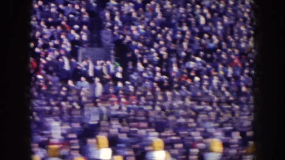Nebraska 1957 football scene with packed stands, sideline activity, and yellow helmets visible near the lower edge.