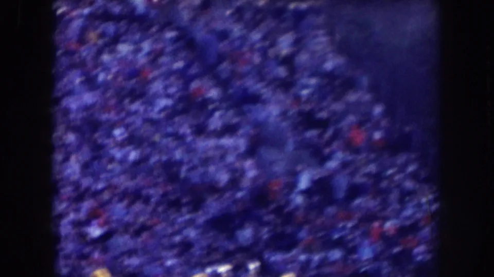 Grainy 1957 Nebraska stadium crowd frame with dense spectators, dark shapes, and scattered red and white highlights.
