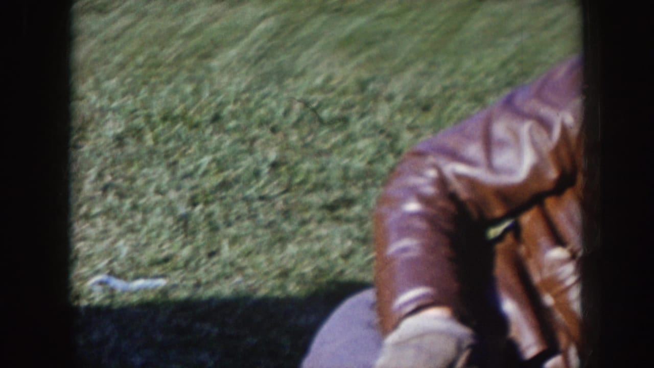 Blurred spectator in a brown leather jacket near grass and dark film-frame edges.