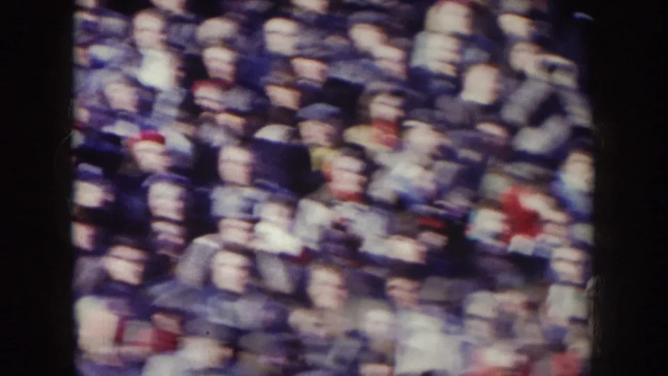1957 Nebraska frame showing rows of spectators packed into bleachers, with faces and clothing softened by grain, focus, and camera movement.