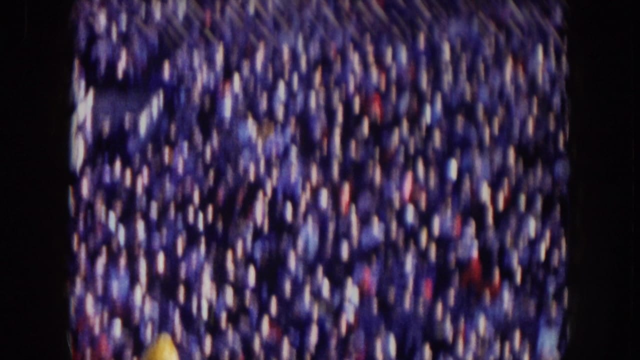 Soft, motion-blurred 1957 Nebraska crowd view with scattered yellow clothing among spectators.