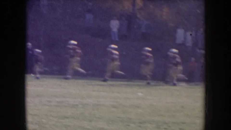 1957 Nebraska 8mm frame showing football players in light uniforms and white helmets jogging across a grassy practice field, with distant bystanders behind them.