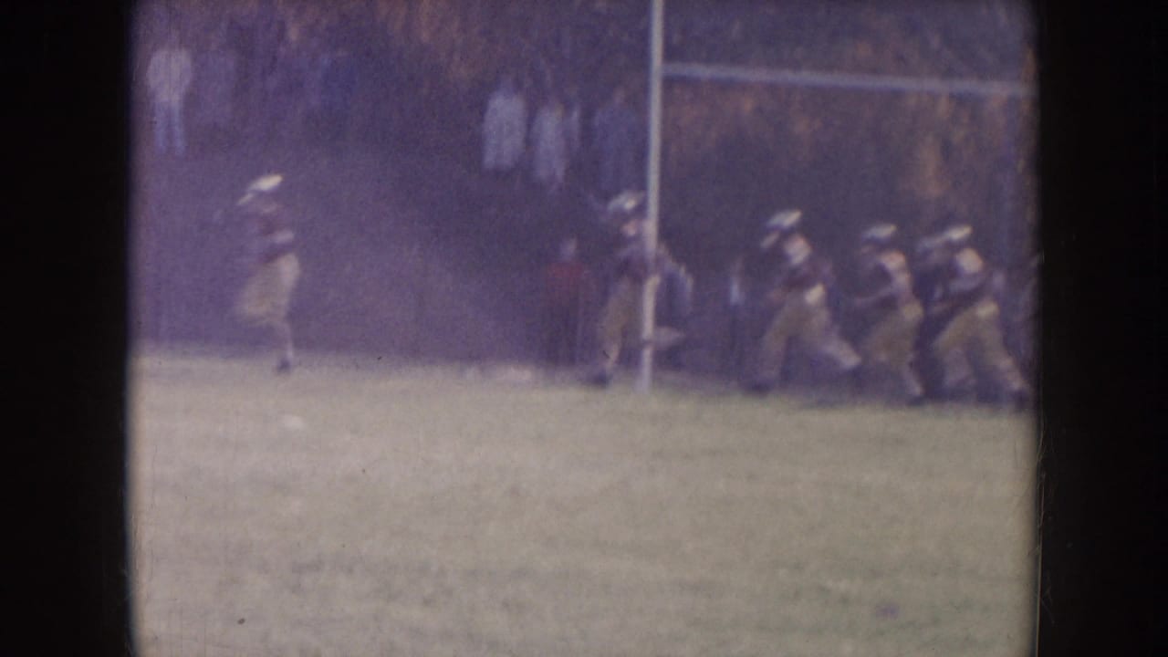 Football players run across a green field near a goalpost.