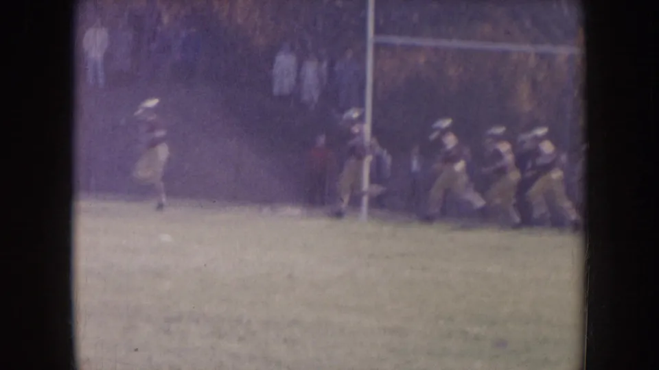 1957 Nebraska home-movie frame showing football players in vintage uniforms running across a green field near a goalpost.