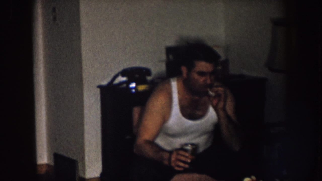 In a Nebraska 1957 home-movie frame, a man in a white tank top smokes indoors while holding a drink near dark furniture.