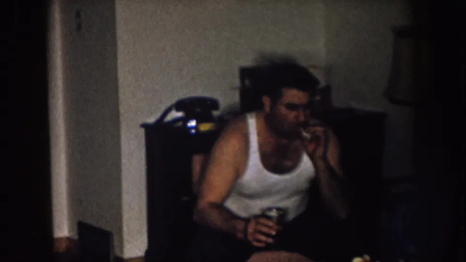 In a Nebraska 1957 home-movie frame, a man in a white tank top smokes indoors while holding a drink near dark furniture.