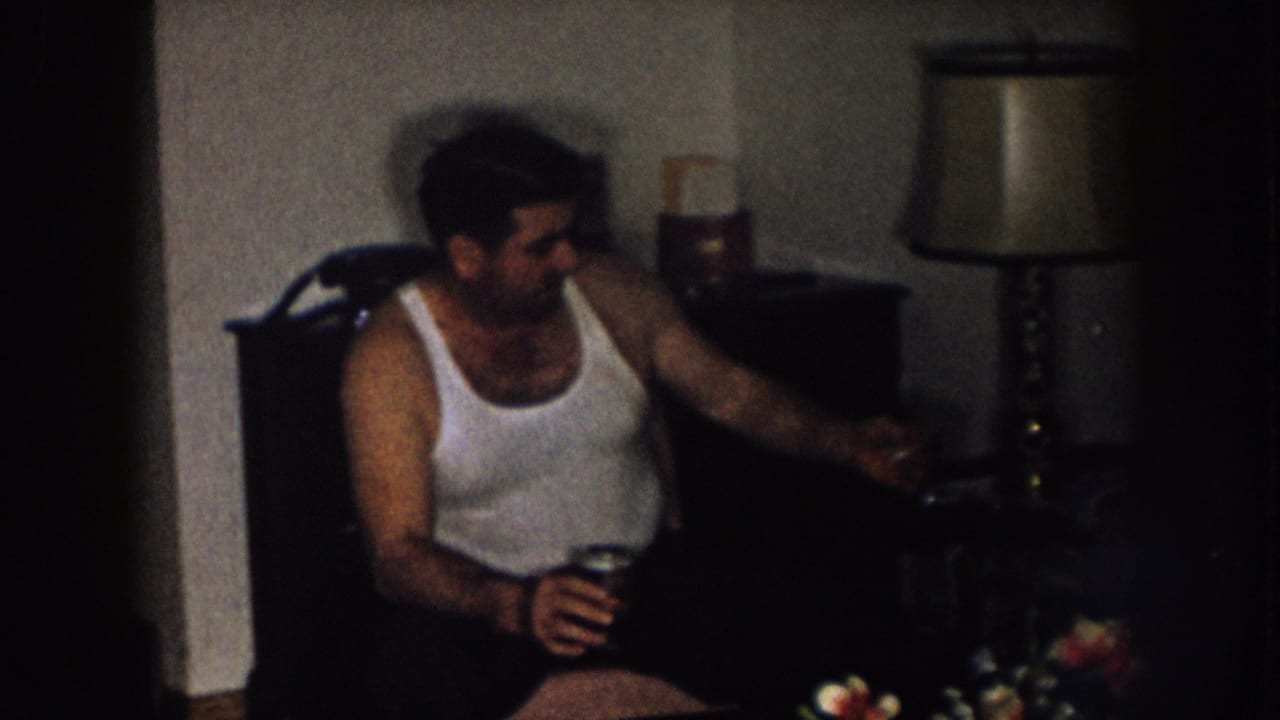 Man in a white tank top reaches toward a side table while holding a drink.