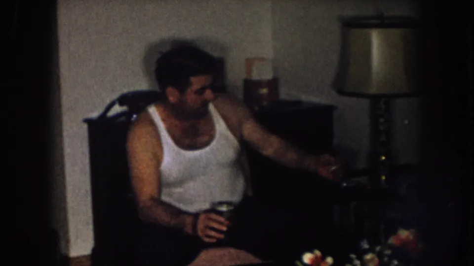 The man sits in the living room with a drink, reaching toward furniture beside a table lamp and household objects.