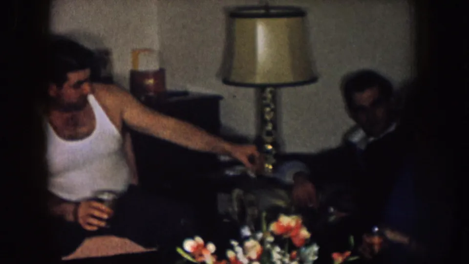 Two adult men are visible in the dim Nebraska living room, with one holding a drink near a lamp and floral arrangement.