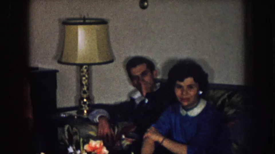 A woman in a blue dress with a white collar sits beside a man on a couch in a 1957 Nebraska living-room scene.