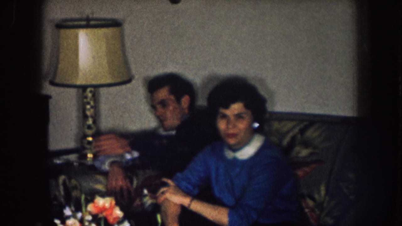 1957 Nebraska home-movie frame showing a woman in a blue sweater and a man seated on a floral couch near a table lamp.