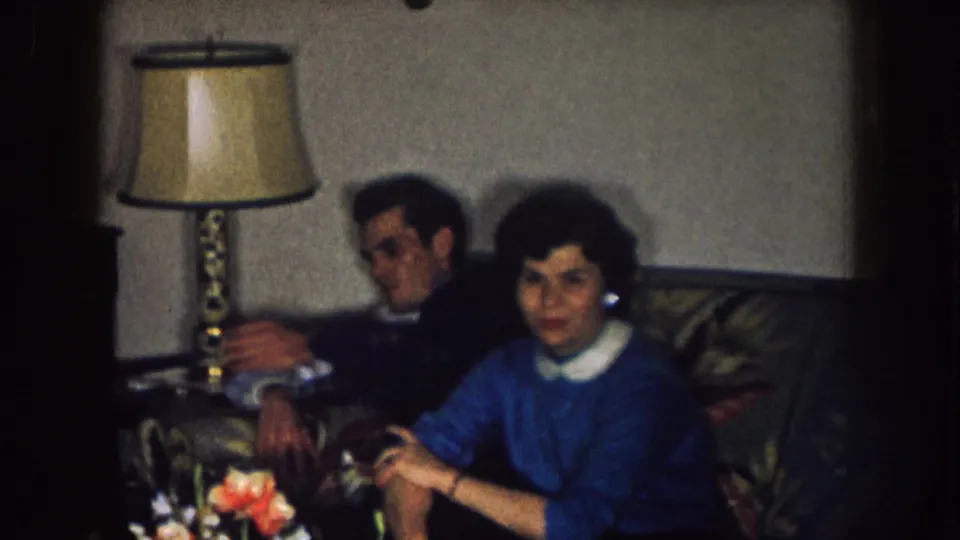 1957 Nebraska home-movie frame showing a woman in a blue sweater and a man seated on a floral couch near a table lamp.