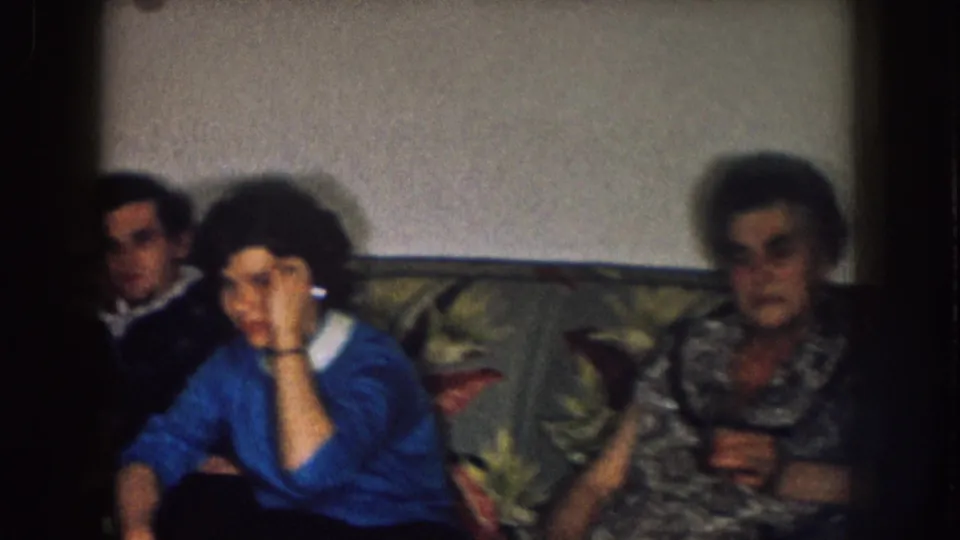 1957 Nebraska home-movie frame with adults seated on a floral couch in a private living room, softly lit by a lamp.