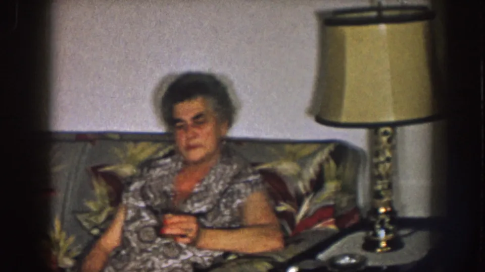 1957 Nebraska home-movie frame showing an older woman seated on a floral couch near a beige-shaded lamp in a living room.