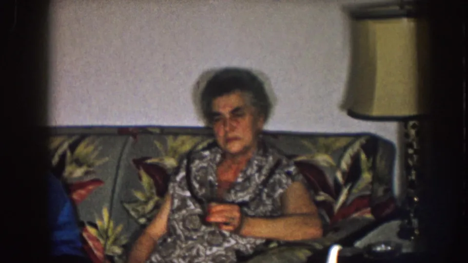 1957 Nebraska home-movie frame of an older woman on a floral couch, with part of another person visible at the left edge.
