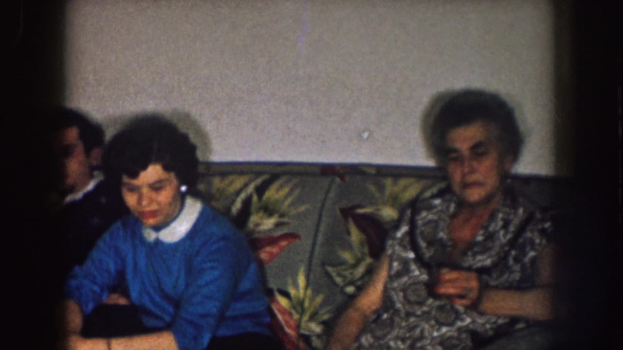 1957 Nebraska home-movie frame showing adults seated on a floral couch in a dim private living room, including women with downcast or stern expressions.
