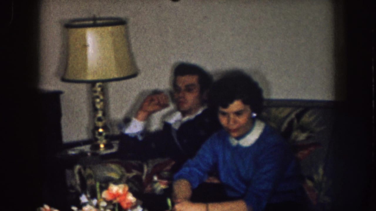 A man and woman sit on a couch near a table lamp while the woman looks down at something in her lap.