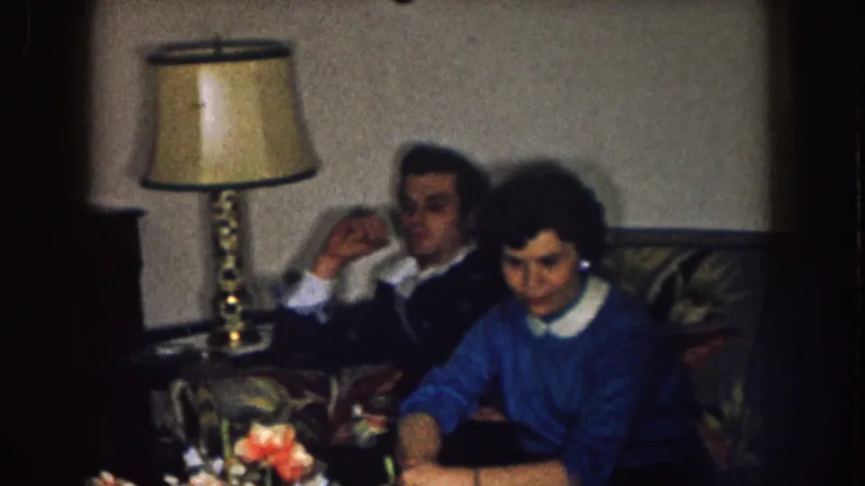 1957 Nebraska interior frame with adults seated on a floral couch beside a table lamp, the mood appearing quiet and uneasy.