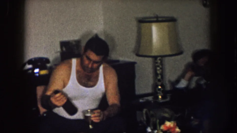 1957 Nebraska home-movie frame showing a man in a white tank top pouring a drink from a bottle, with seated adults blurred behind him.