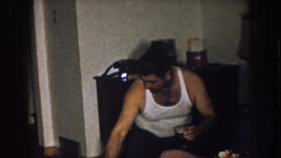 1957 Nebraska interior frame showing a man holding a drink and leaning forward near a doorway and dark furniture in a private living room.