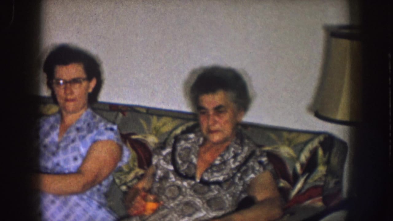 Two women sit on a patterned couch in a dim living room.