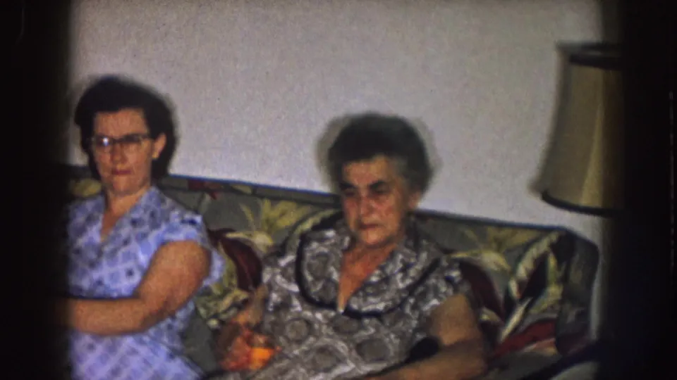Two adult women sit on a patterned couch in a Nebraska living room, 1957, with one holding a drink.