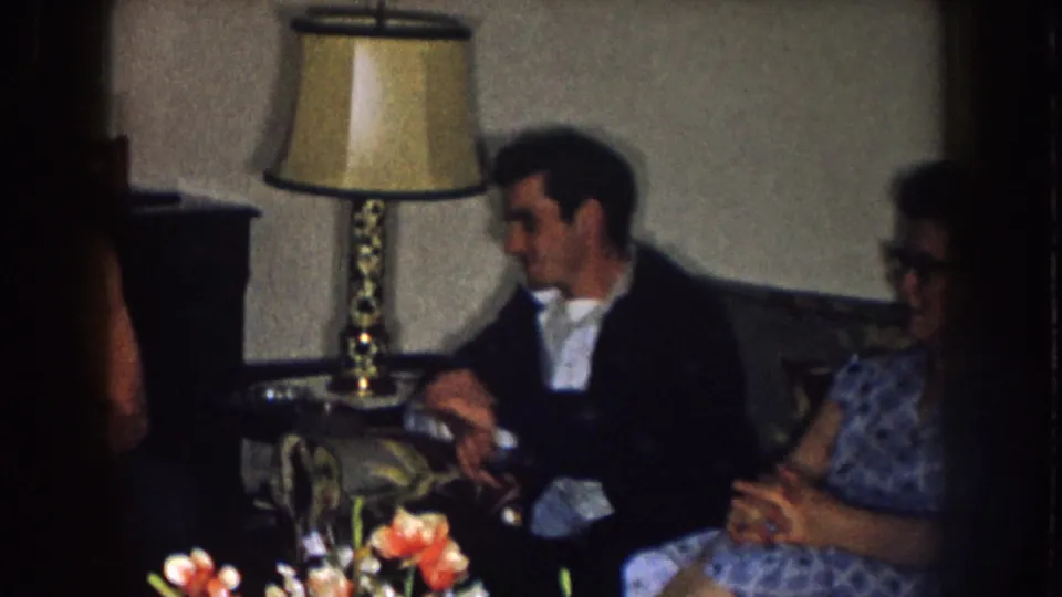 A man in a dark jacket sits beside a woman on a couch in a Nebraska living room, with a table lamp and flowers in front.