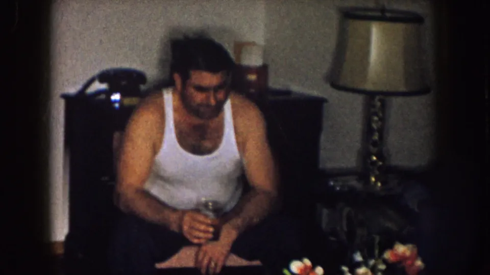 A seated man in a white tank top holds a glass in a 1957 Nebraska living room, with a lamp and flowers nearby.
