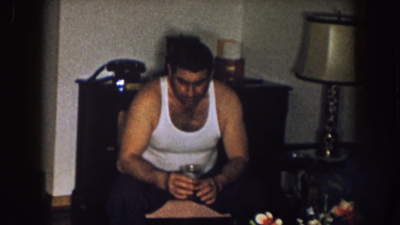 In a 1957 Nebraska living room, a man in a white tank top sits holding a glass beside a rotary phone, lamp, and table.