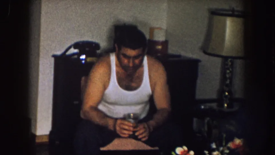 In a 1957 Nebraska living room, a man in a white tank top sits holding a glass beside a rotary phone, lamp, and table.