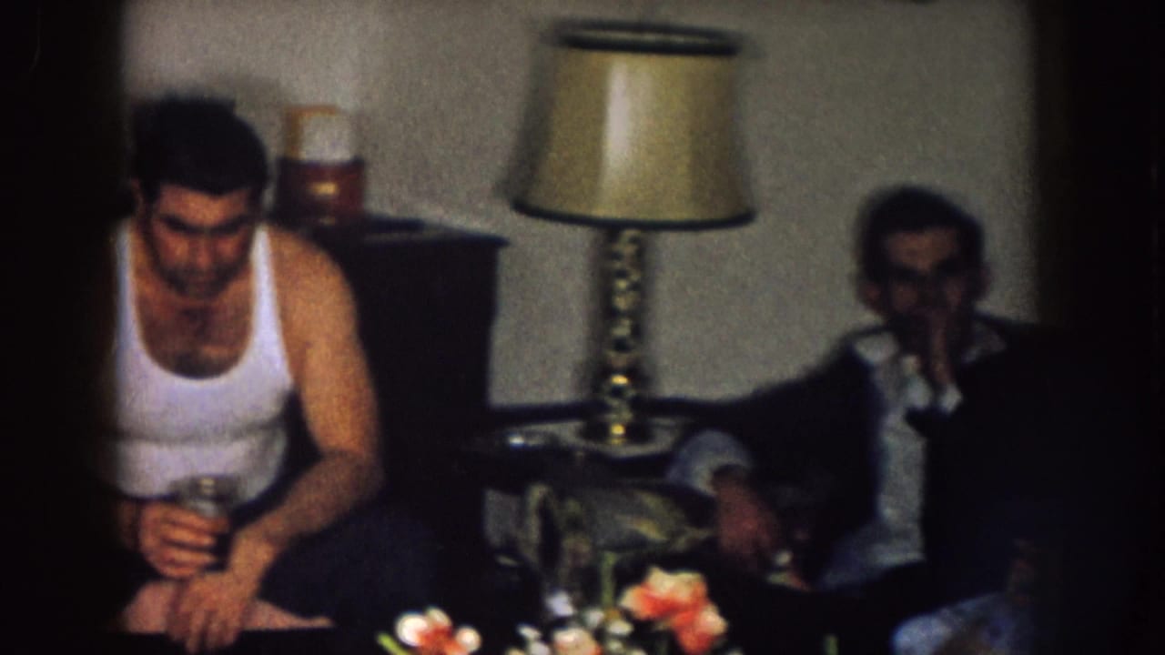 Man holding a glass sits beside another adult on a couch in a dim living room.