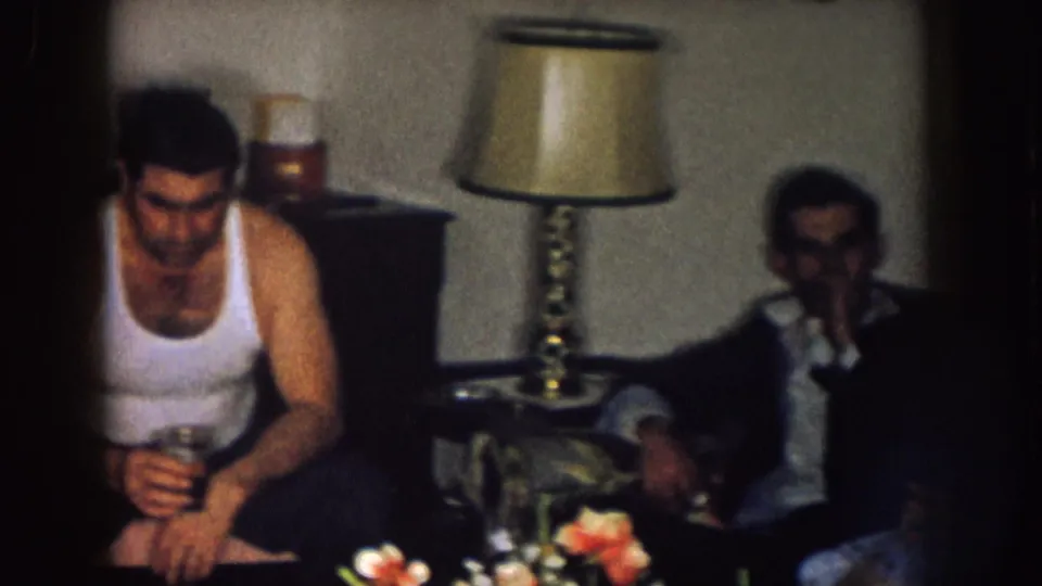 A Nebraska home-movie frame shows a seated man with a glass while another adult sits nearby under a table lamp.